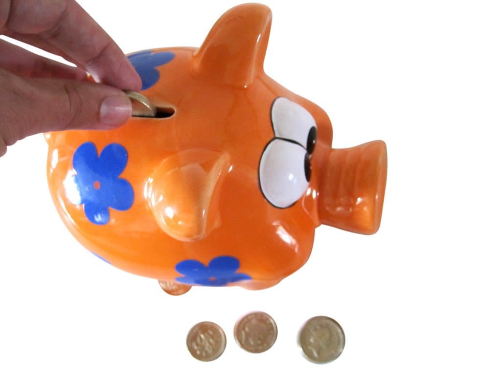 A hand dropping a coin into an orange piggy bank, with more coins below
