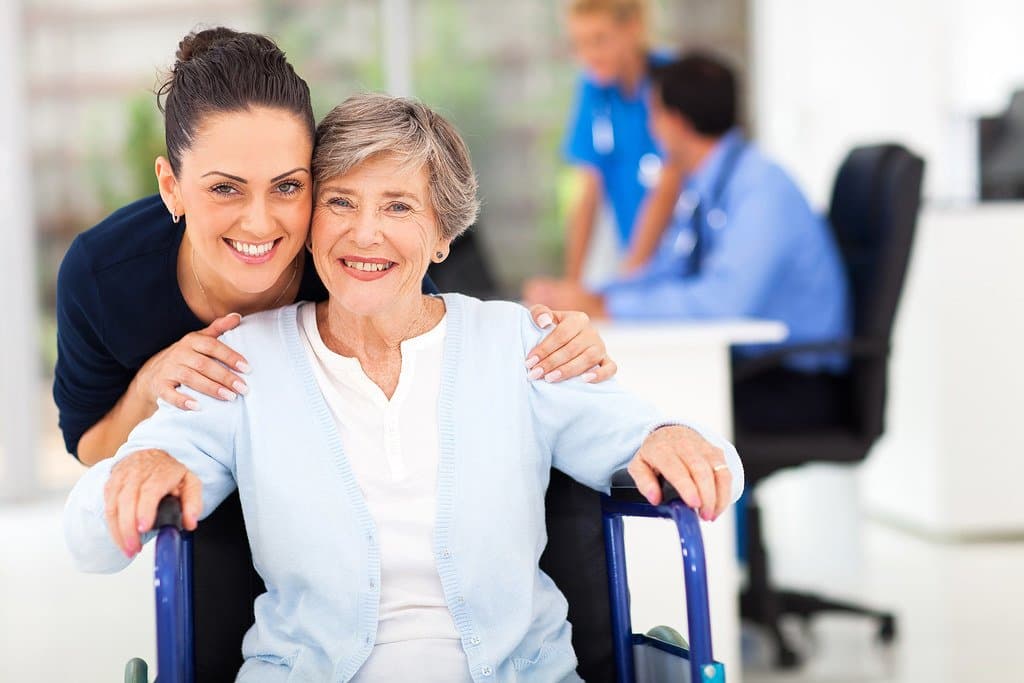 An adult daughter hugging her smiling mother, who sits in a wheelchair at a clinic