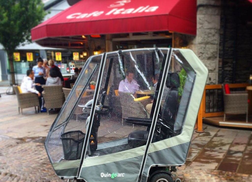An enclosed mobility scooter with a clear canopy parked outside a café on a cobblestone street