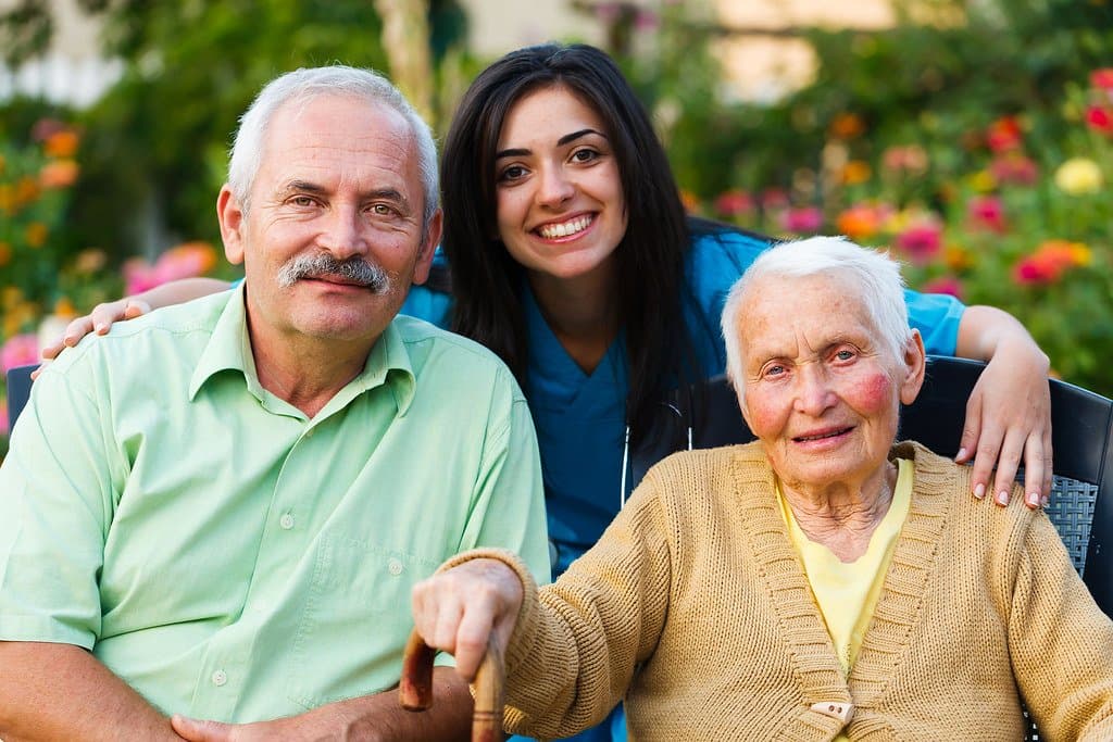 A man and a young woman smiling beside an elderly woman with a cane in a garden