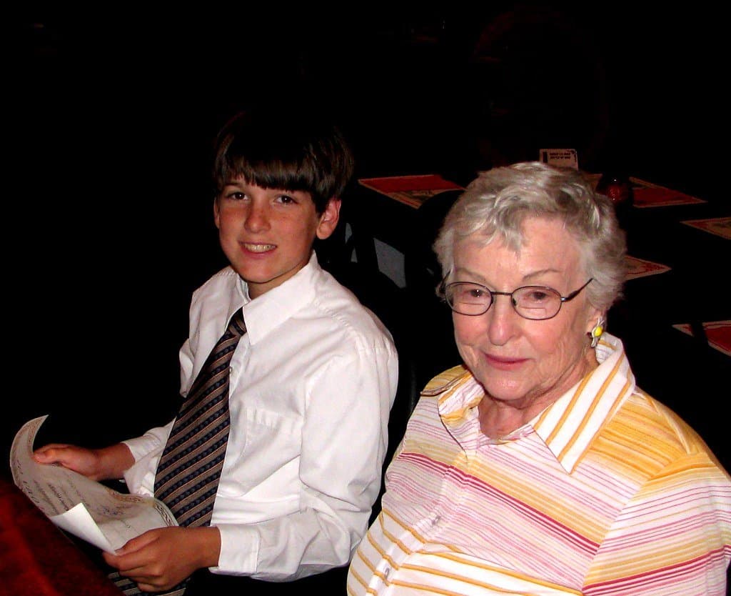 A grandmother in glasses sitting beside her teenage grandson in a shirt and tie