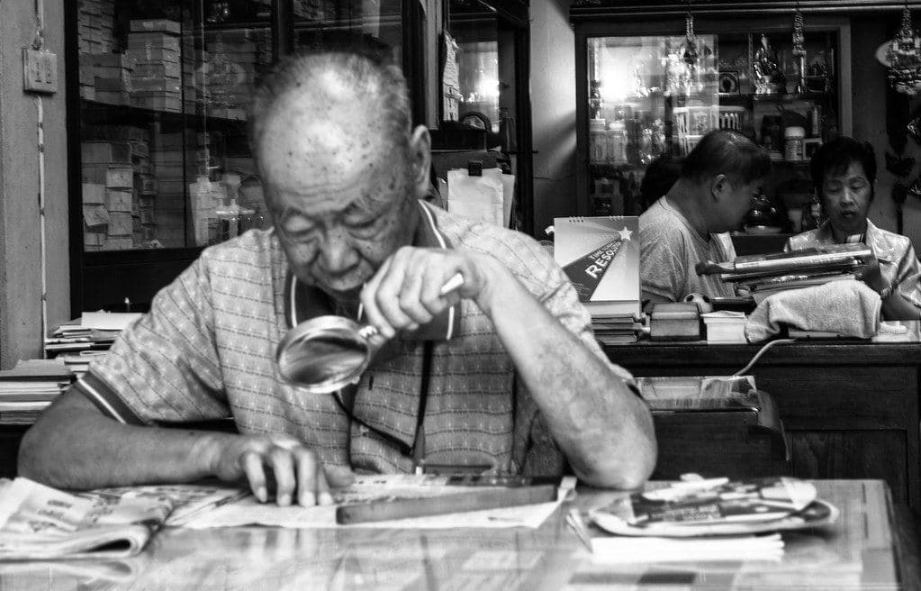 An older man reading a newspaper through a handheld magnifying glass at a table