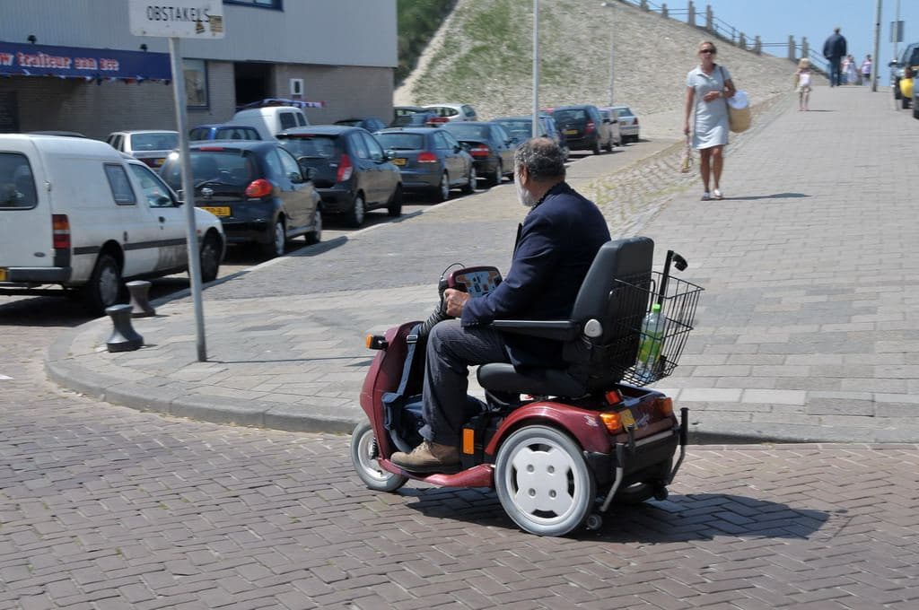 A man in a navy jacket riding a red four-wheel mobility scooter along a paved waterfront street
